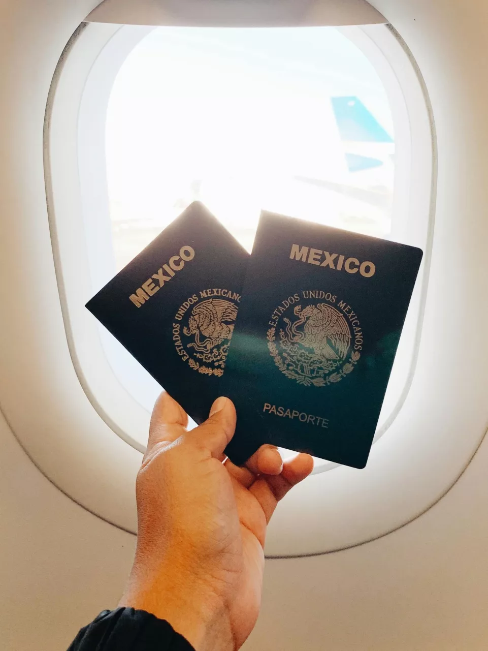 mano sosteniendo dos pasaportes mexicanos en frente de una ventana de avión.